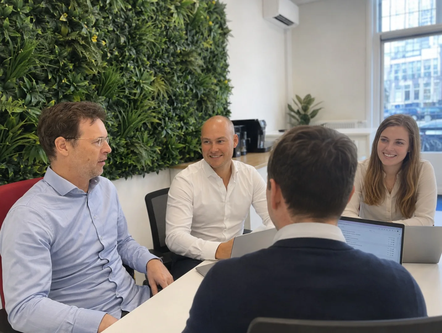 Four Agentancy team members talking at the meeting table in the office, laptops open and a green plant wall in the background.